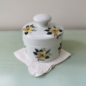 May include: White ceramic jar with a lid, decorated with yellow roses and green leaves. The jar is sitting on a white doily.