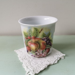 Peut inclure: Une tasse en céramique blanche avec un motif de fruits colorés représentant des pommes, des raisins et des baies. La tasse est posée sur une dentelle blanche crochetée.