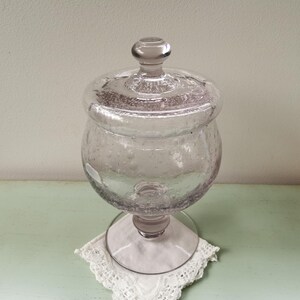 Peut inclure: Un pot à bonbons en verre transparent avec un couvercle et un piédestal. Le pot a une surface texturée, bulleuse et une teinte légèrement violette.