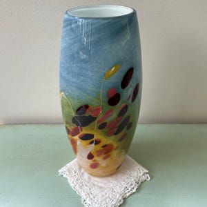 May include: A tall, cylindrical glass vase with a white rim. The vase has a blue, green, and yellow background with abstract red, black, and brown spots.
