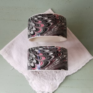May include: Two ceramic napkin rings with a marbled design in black, white, and pink.