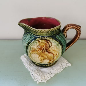 Puede incluir: Una jarra de cerámica verde y marrón con un diseño de león. La jarra tiene un interior rojo y un asa marrón.