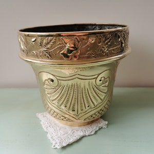 Puede incluir: Un macetero de latón de color dorado con un diseño ornamentado. El macetero tiene un diseño de conchas en la parte inferior y un diseño floral alrededor de la parte superior.
