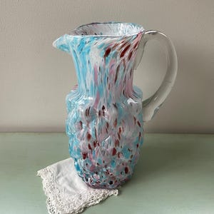 May include: A colorful glass pitcher with a clear handle. The pitcher is decorated with blue, pink, and maroon speckles. A white lace doily rests beneath the pitcher. This decorative pitcher is a unique addition to any home decor.