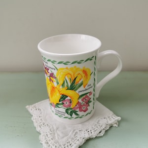Peut inclure: Une tasse en céramique blanche avec un motif floral représentant des iris jaunes, des fleurs roses et des feuilles vertes. La tasse a une anse et le texte "Iris pseudacorus" et "Cardamine pratensis" est imprimé sur le côté.