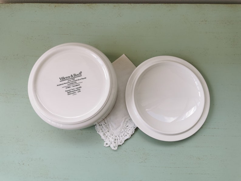 May include: White ceramic lidded dish with the text "Villeroy & Boch" and "Anmut" on the bottom. The dish is on a light green surface.