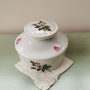 May include: White porcelain jar with a lid, decorated with pink roses and green leaves. The jar is sitting on a white lace doily.