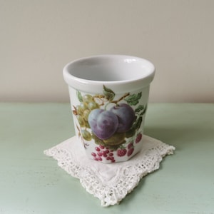 May include: A white ceramic cup with a fruit design. The cup features plums, grapes, and raspberries in shades of purple, green, and red. The cup is sitting on a white crocheted doily.