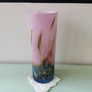 May include: A tall, cylindrical glass vase with a pink and blue marbled design. The vase has a light pink base with streaks of gold and brown, transitioning to a dark blue base with swirls of green and gold.