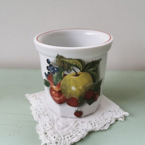 Peut inclure: Pot en céramique blanc avec un motif de fruits. Le pot présente une illustration colorée d'une pomme, de fraises et de myrtilles. Le bord a un fin liseré rouge. Le pot repose sur un napperon en dentelle blanche.