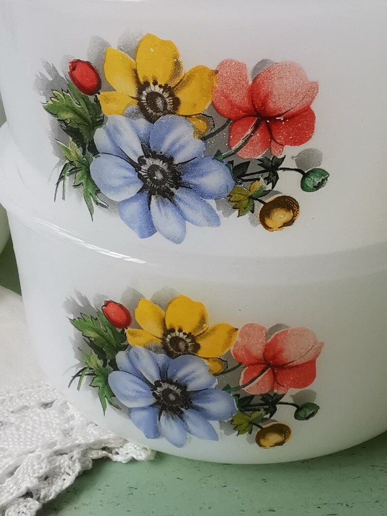 May include: Two white ceramic bowls with a floral design. The design features yellow, blue, and red flowers with green leaves. The bowls are stacked on top of each other.