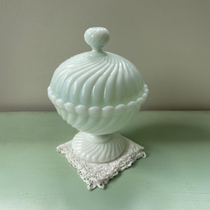 Puede incluir: Un plat de dulces vintage de vidrio de leche azul claro con un diseño de espiral y una tapa. El plato está sobre una servilleta de encaje blanca.