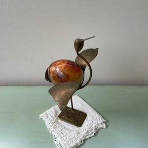 Puede incluir: Una escultura de pájaro vintage con un cuerpo de mármol en tonos naranja y marrón, sostenida por alas de latón y un cuello delgado. La escultura descansa sobre una base cuadrada de latón, encima de un mantelito blanco con bordes de encaje. El pico del pájaro es puntiagudo.
