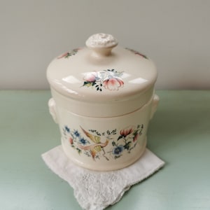Peut inclure: Un pot en céramique blanche avec un couvercle, décoré d'un motif floral dans des tons de rose, bleu et vert. Le pot a deux petites poignées sur les côtés.