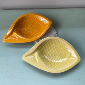 May include: Two ceramic fish-shaped serving dishes. One is a bright yellow-orange, the other a pale yellow. Both have textured scale patterns and a small fin detail. The dishes are on a light blue surface.