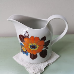 May include: White ceramic jug with a floral design in orange, yellow, blue, and brown. The jug has a rounded body and a handle.