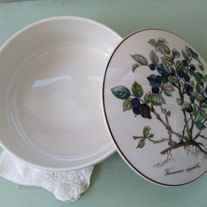 May include: White ceramic serving bowl with a lid. The lid features a botanical illustration of a blueberry bush with green leaves and blue berries. The text "Vaccinium myrtillus" is printed on the lid.