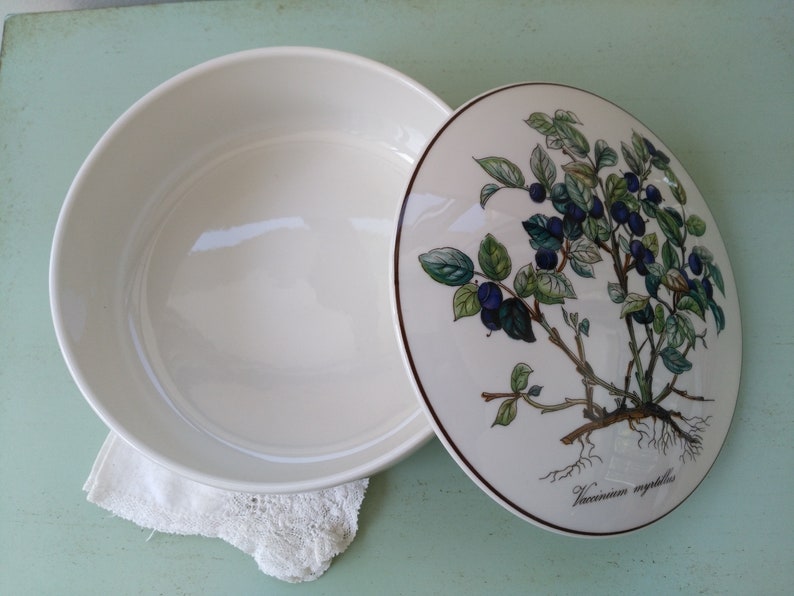May include: White ceramic serving bowl with a lid. The lid features a botanical illustration of a blueberry bush with green leaves and blue berries. The text "Vaccinium myrtillus" is printed on the lid.