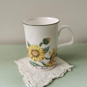 Könnte beinhalten: Weiße Keramiktasse mit grünem Rand und einem Blumendesign mit gelben Gänseblümchen und grünen Blättern. Die Tasse hat einen Henkel und den Text "Corn Marigold" auf dem Boden.