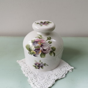 Puede incluir: Jarrón de cerámica blanca con un diseño floral. El jarrón tiene una tapa con un diseño floral. El jarrón está sentado sobre una servilleta blanca.