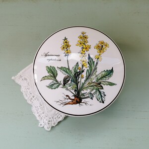 May include: A white ceramic lidded box with a botanical illustration of a yellow flower with green leaves and the text "Agrimonia eupatoria".