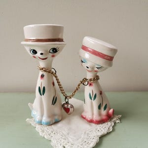 Könnte beinhalten: Zwei weiße Keramik-Katzenfiguren, die Hüte tragen und eine goldene Kette mit einem Herz-Anhänger halten. Die Katze links trägt einen weißen Hut mit einem braunen Band. Die Katze rechts trägt einen weißen Hut mit einem rosa Band. Beide Katzen haben blaue Augen und sind mit grünen und roten Blumenmustern verziert.