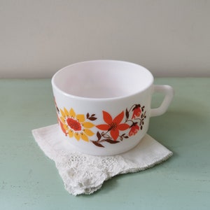 Op de afbeelding: Witte keramische mok met een bloemenontwerp in tinten oranje, geel en bruin. De bloemen zijn gestileerd en lijken op zonnebloemen en andere bloemen.