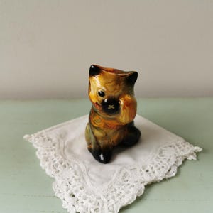 Op de afbeelding: Een klein keramiek beeldje van een kat met bruine, zwarte en gele markeringen. De kat zit met zijn poten ingetrokken en zijn hoofd naar beneden gekanteld. Het beeldje staat op een wit kantje.