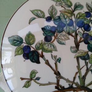 May include: A white ceramic plate with a hand-painted design of a blueberry bush with green leaves and blue berries. The plate has a gold rim.