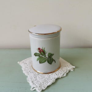 May include: White ceramic jar with a gold trim and a single rose design. The jar has a lid and is sitting on a white doily.