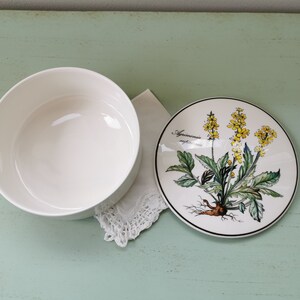 May include: A white ceramic bowl with a floral design on the lid. The lid features a yellow flower with green leaves and the text "Agrimonia eupatoria".