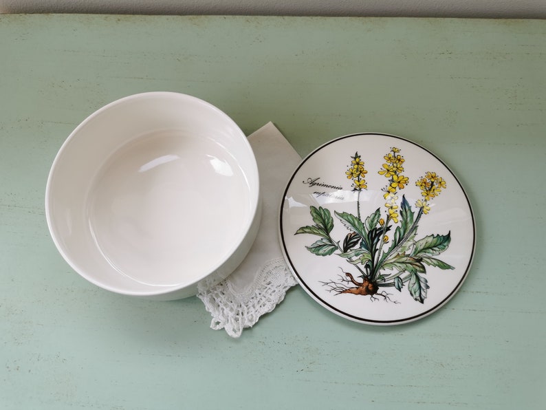 May include: A white ceramic bowl with a floral design on the lid. The lid features a yellow flower with green leaves and the text "Agrimonia eupatoria".