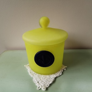 Può includere: Un barattolo di vetro giallo smerigliato con un coperchio e un sigillo blu. Il barattolo è su un tovagliolo bianco.