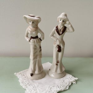 Peut inclure: Deux figurines en porcelaine de personnes portant des chapeaux et des robes. Les figurines sont blanches avec des accents bruns. La femme de gauche porte un chapeau blanc avec un bandeau brun et une robe blanche avec des garnitures brunes. La femme de droite porte un chapeau blanc avec un bandeau brun et une robe blanche avec des garnitures brunes. Les figurines sont debout sur une dentelle blanche.