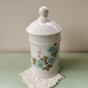 May include: White ceramic canister with a floral design featuring blue and pink flowers. The canister has a lid and is perfect for storing sugar, tea, or other kitchen staples.