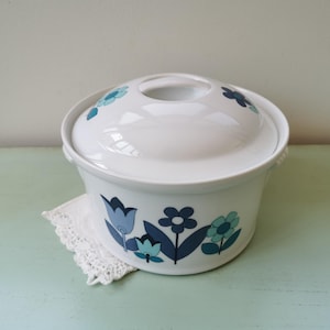 Peut inclure: Plat à casserole en céramique blanche avec couvercle, orné de motifs floraux bleus et turquoise. Le plat repose sur un napperon en dentelle blanche. Le couvercle a une ouverture circulaire. Article de cuisine vintage.