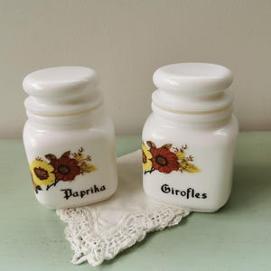 Op de afbeelding: Twee witte keramische kruidenpotten met bloemmotieven en deksels. De potten zijn gelabeld met "Paprika" en "Girofles".