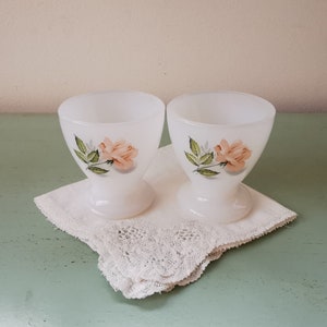 Könnte beinhalten: Zwei weiße Keramik-Eierbecher mit einem Design aus rosa Rosen und grünen Blättern. Die Becher stehen auf einer weißen Spitzenunterlage.