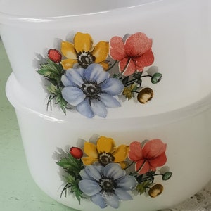 May include: Two white ceramic bowls with floral designs. The bowls feature a cluster of blue, yellow, and red flowers with green leaves. The bowls are stacked on top of each other.