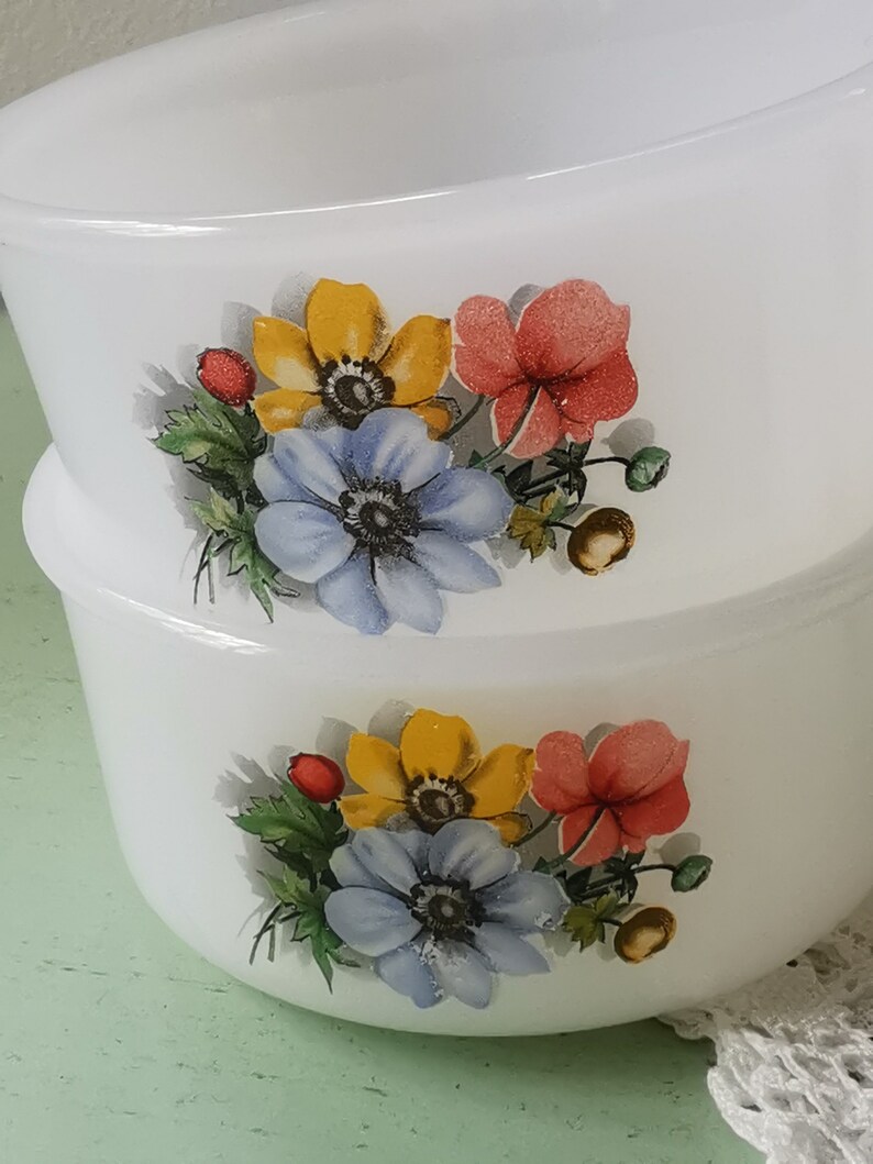May include: Two white ceramic bowls with floral designs. The bowls feature a cluster of blue, yellow, and red flowers with green leaves. The bowls are stacked on top of each other.