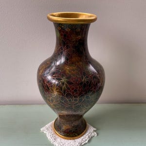 Pode incluir: Um vaso decorativo com um padrão floral em tons de castanho, vermelho e verde. O vaso tem uma borda e base douradas. É colocado sobre uma toalha de renda branca.