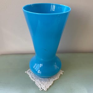 Puede incluir: Un jarrón de vidrio azul brillante con una base redondeada y una abertura acampanada. El jarrón está sobre un encaje de ganchillo blanco.