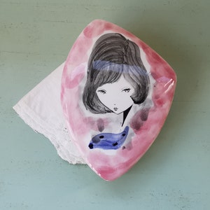 Può includere: Un piatto in ceramica rosa con un'illustrazione stilizzata di una donna con i capelli neri e una sciarpa blu. Il piatto ha la forma di un diamante.