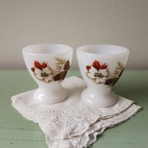 Puede incluir: Dos hueveras de cerámica blanca con diseños florales. Las hueveras tienen un fondo blanco con flores marrones y rojas y hojas verdes.