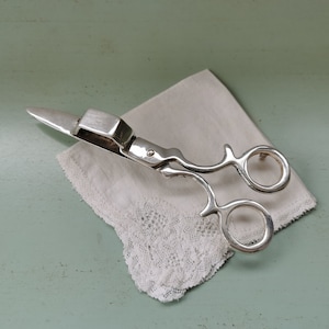 May include: Silver-toned scissor-shaped napkin ring with a decorative design. The ring is resting on a white lace handkerchief.