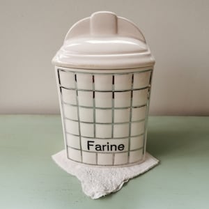 May include: White ceramic canister with a grid pattern and the word "Farine" in black lettering. The canister has a lid and is sitting on a white doily.