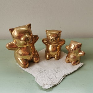 Op de afbeelding: Drie gouden messing beeldjes van beren, elk met een andere maat. De grootste beer staat op zijn achterpoten met zijn voorpoten omhoog. De middelste beer staat op zijn achterpoten met zijn voorpoten langs zijn zijde. De kleinste beer zit op zijn achterpoten met zijn voorpoten langs zijn zijde.
