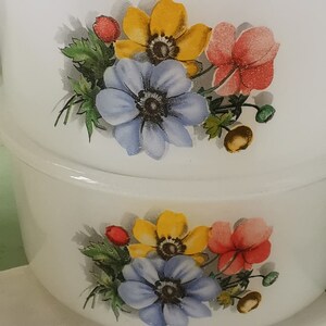 May include: Two white ceramic bowls with floral designs. The bowls feature yellow, blue, and red flowers with green leaves. The bowls are stacked on top of each other.