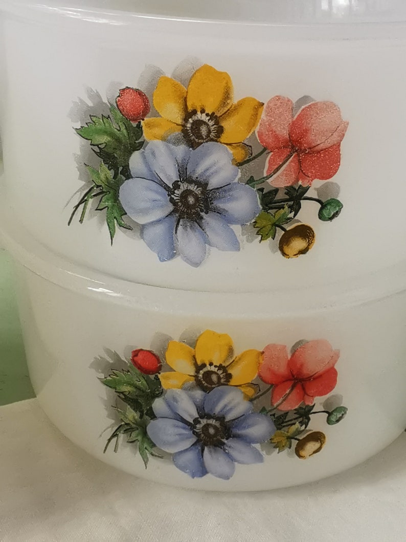May include: Two white ceramic bowls with floral designs. The bowls feature yellow, blue, and red flowers with green leaves. The bowls are stacked on top of each other.