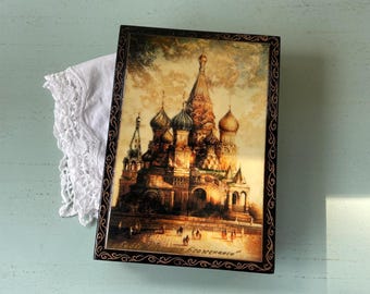 May include: A rectangular, black-framed box with a detailed painting of St. Basil's Cathedral in Moscow. The artwork features warm tones of gold, brown, and red, with intricate architectural details. The box rests on a white lace cloth.